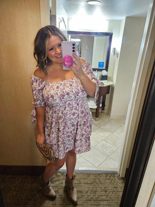 A woman takes a mirror selfie in a hotel bathroom while wearing a short off-the-shoulder floral dress with a purple and white pattern, holding a pink phone case.